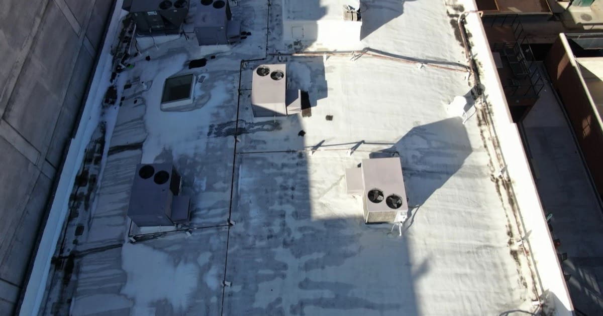 Aerial view of a weathered low-slope commercial roof in Denver, Colorado, showing a worn coating, rooftop HVAC units, and debris collecting along the parapet edges
