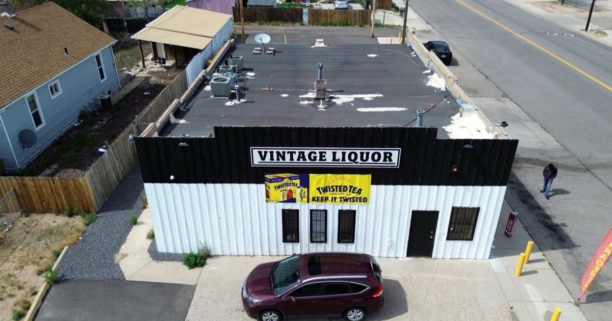 Aerial view of an aging dark asphalt low-slope roof on a small commercial building in Greeley, Colorado, with pale patch repairs spread across the roof field