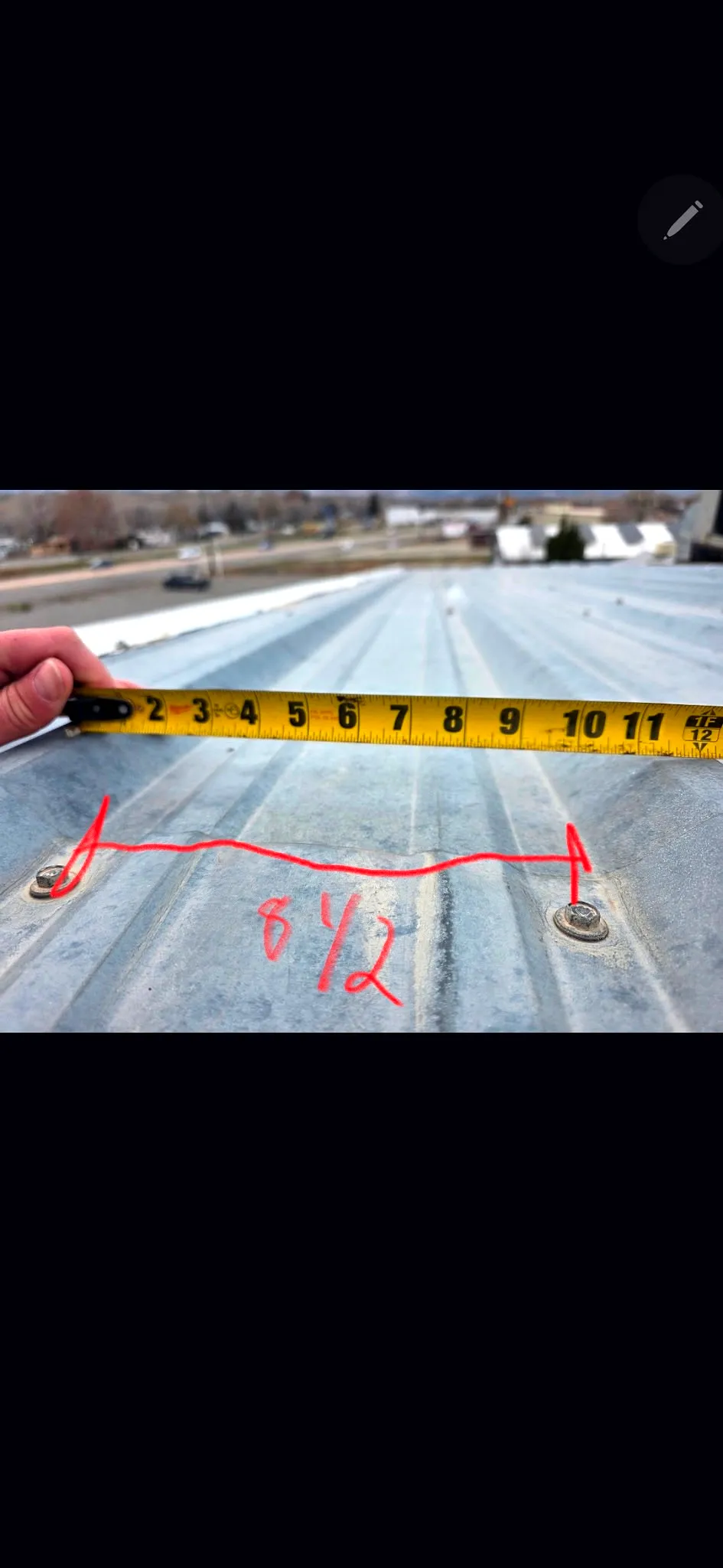 A tape measure laid across ribbed galvanized metal roof panels showing roughly 8-1/2 inch rib spacing, with an exposed fastener marked, on a Grand Junction commercial roof