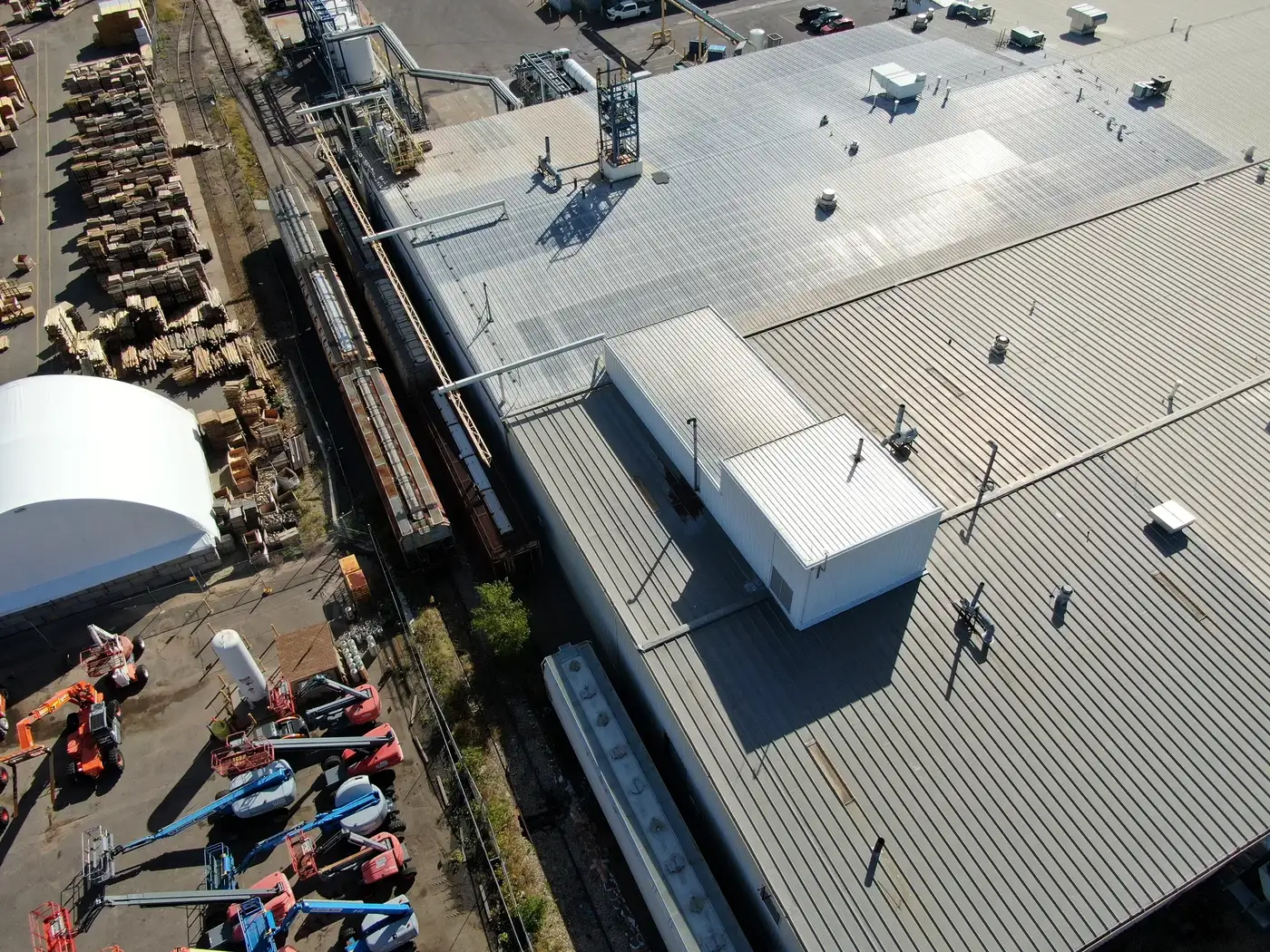 Drone view along the side of an industrial metal-roofed building in Denver, Colorado, with rail cars on the spur, the penthouse, and an equipment yard next door