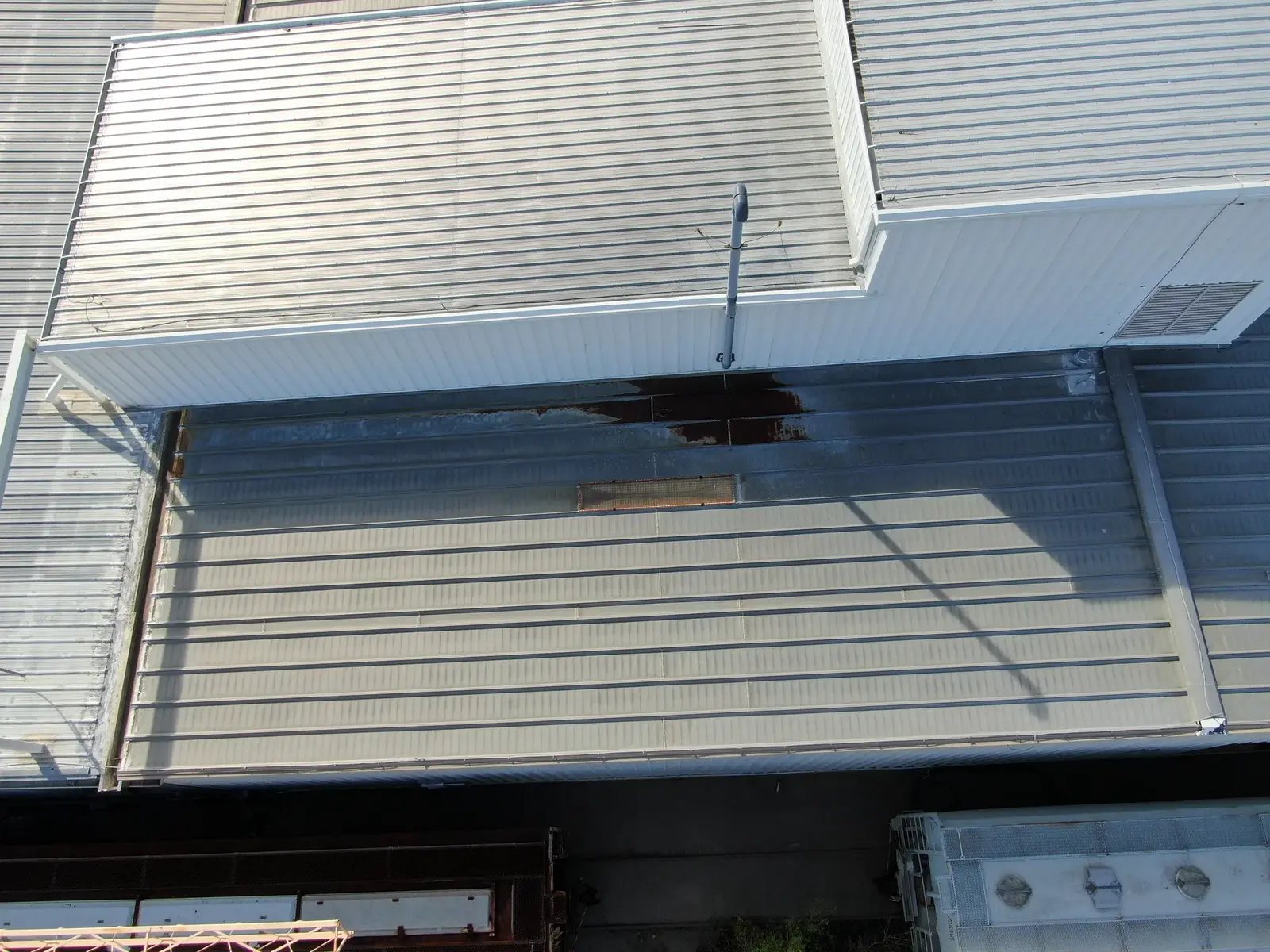 Drone close-up of ribbed metal roof panels beside a white penthouse on an industrial building in Denver, Colorado, with rust staining at the panel ends along the penthouse wall