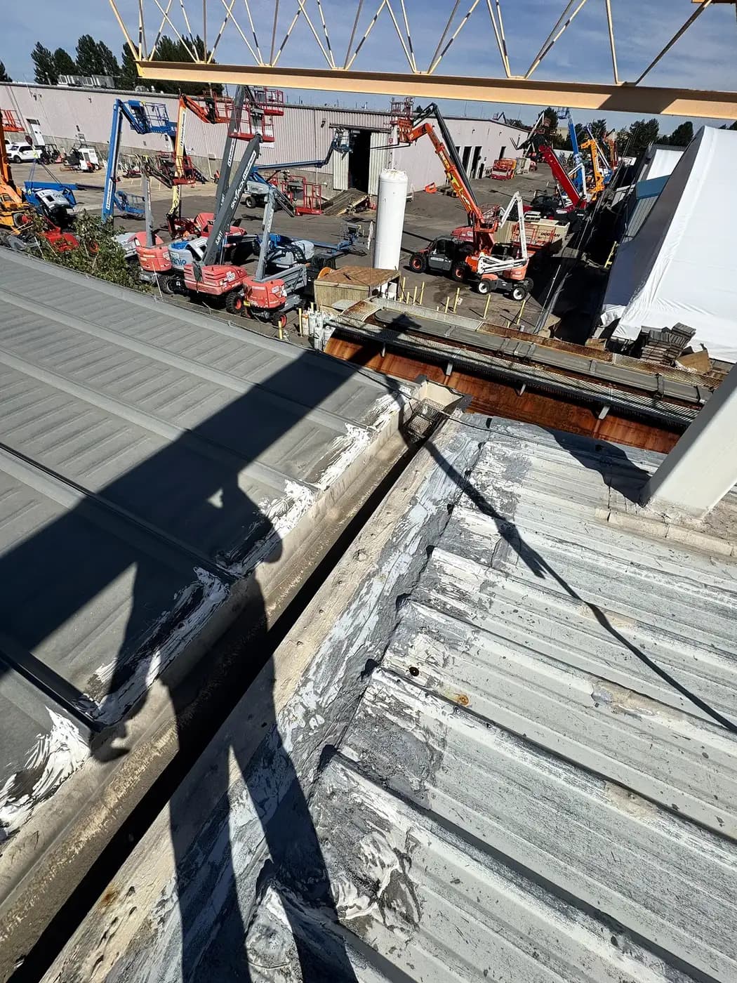 The eave edge of an older ribbed metal roof on an industrial building in Denver, Colorado, heavily patched with white sealant along the gutter line