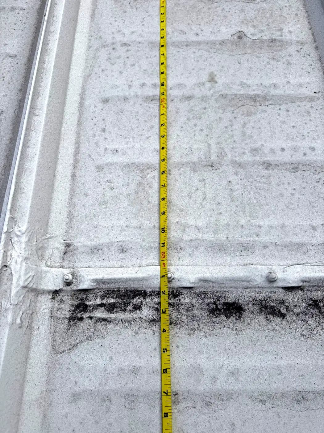 A tape measure laid across a ribbed metal roof panel end lap with exposed fasteners and dark staining on an industrial roof in Denver, Colorado