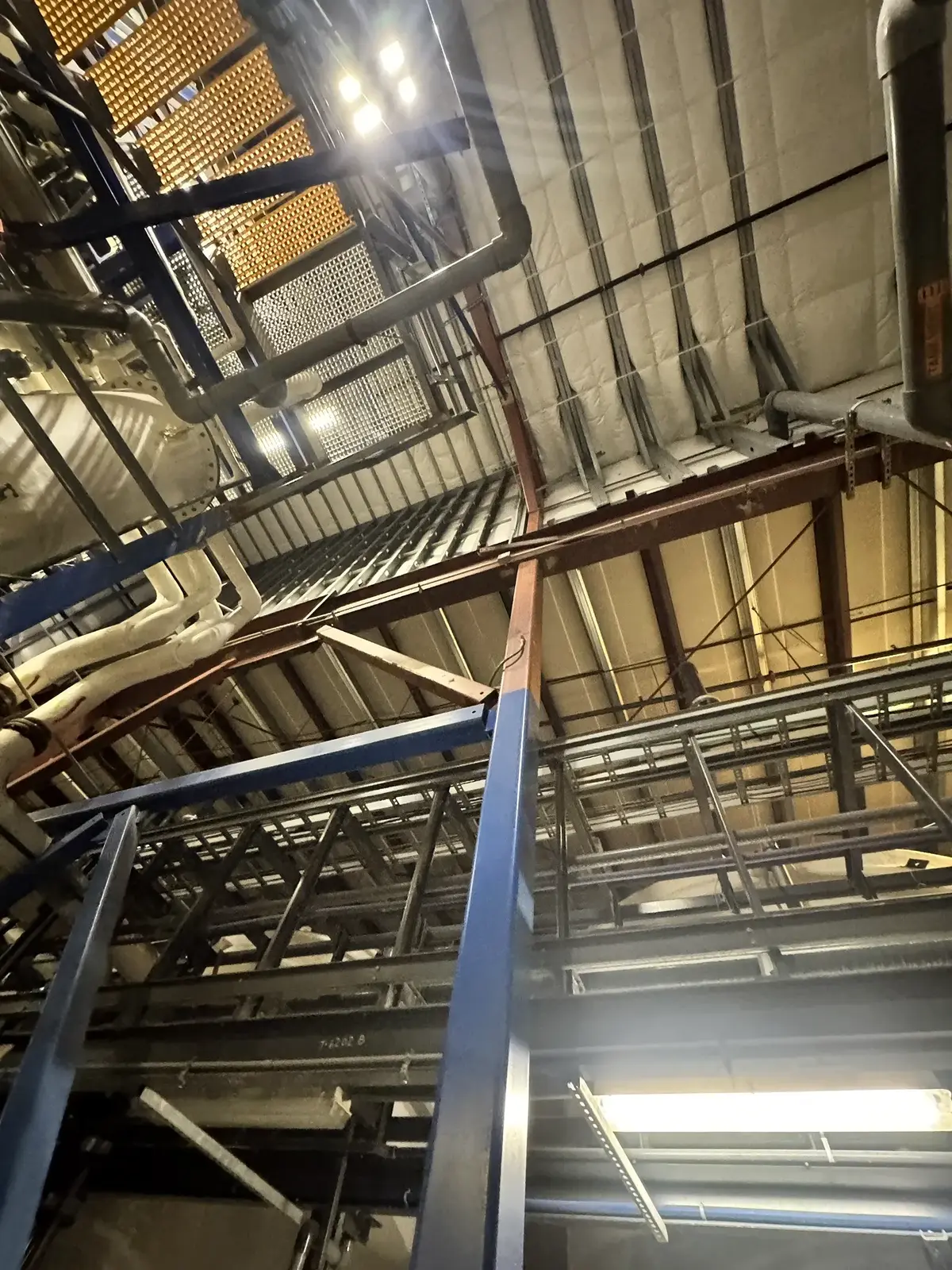 Steel roof framing, purlins, and process piping seen from inside an industrial chemical facility in Denver, Colorado, with light showing through at the roof line