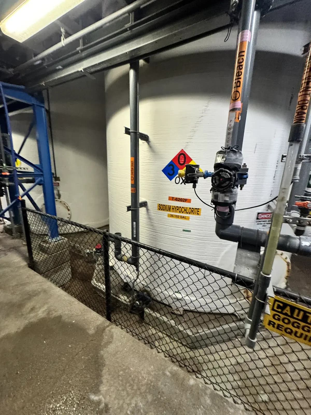 A large white sodium hypochlorite storage vat with hazard placards and piping behind a chain-link guard inside an industrial chemical facility in Denver, Colorado