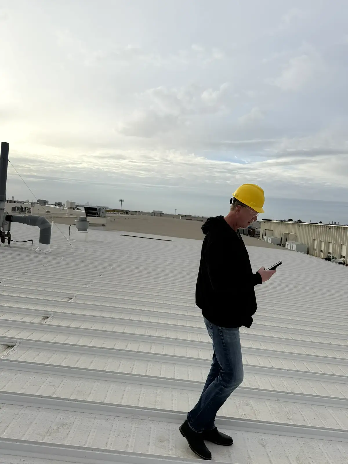 A roofer in a yellow hard hat checking notes on a phone while inspecting a large low-slope metal roof on an industrial building in Denver, Colorado