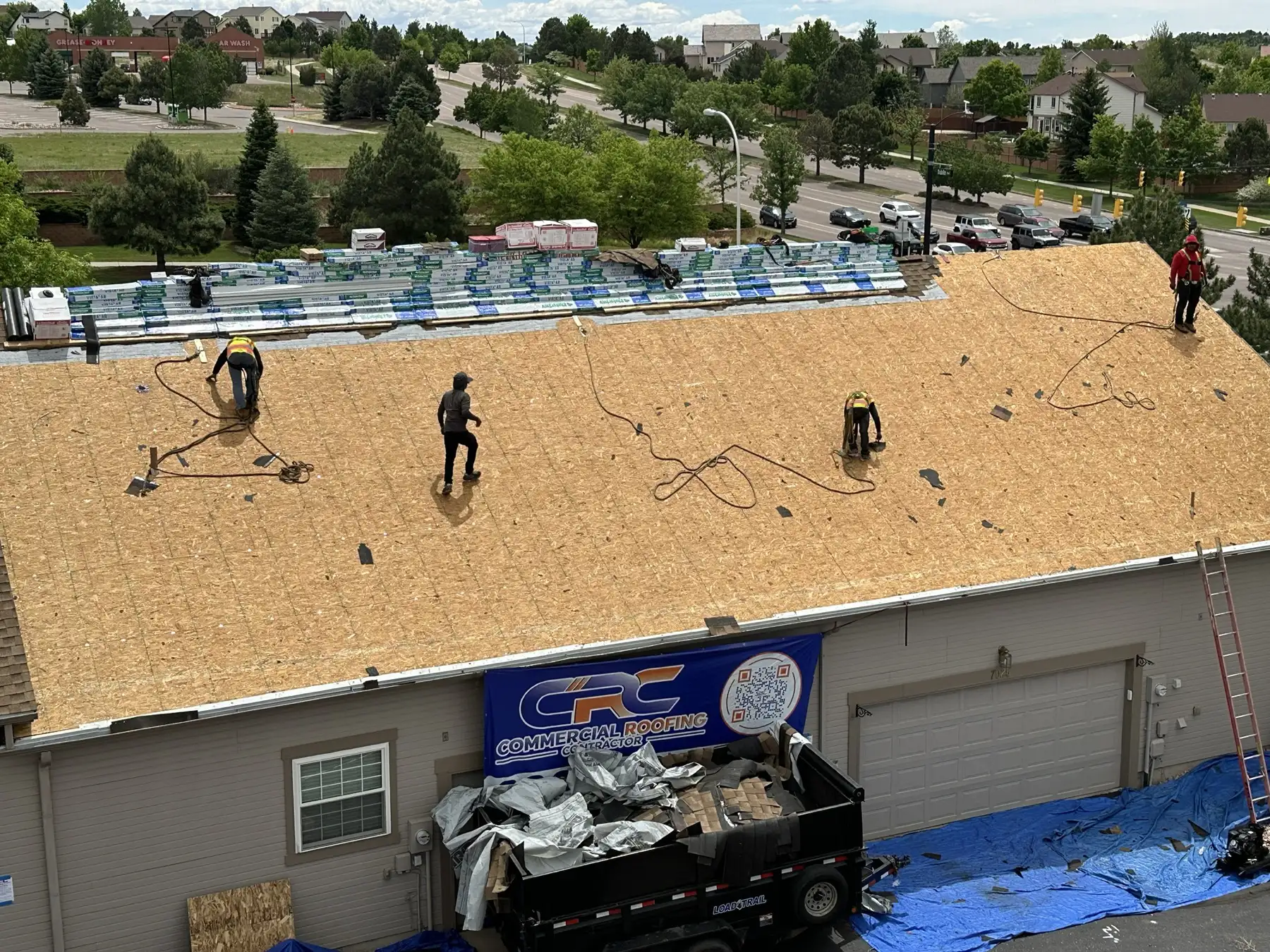 A condominium building in Colorado Springs stripped to bare wood decking with a Commercial Roofing Contractor banner hung on the building and the crew and dump trailer staged below