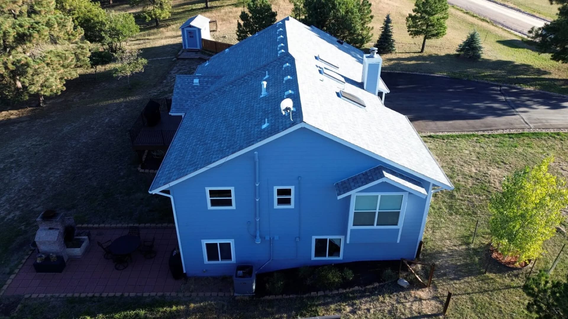 Completed architectural asphalt shingle roof on a two-story blue home on a wooded acreage in Elizabeth, Colorado, in golden-hour light