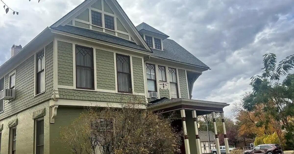 A green historic two-storey home in Grand Junction, Colorado with several distinct roof planes: a main hipped roof, a gabled dormer, a smaller dormer and a separate porch roof